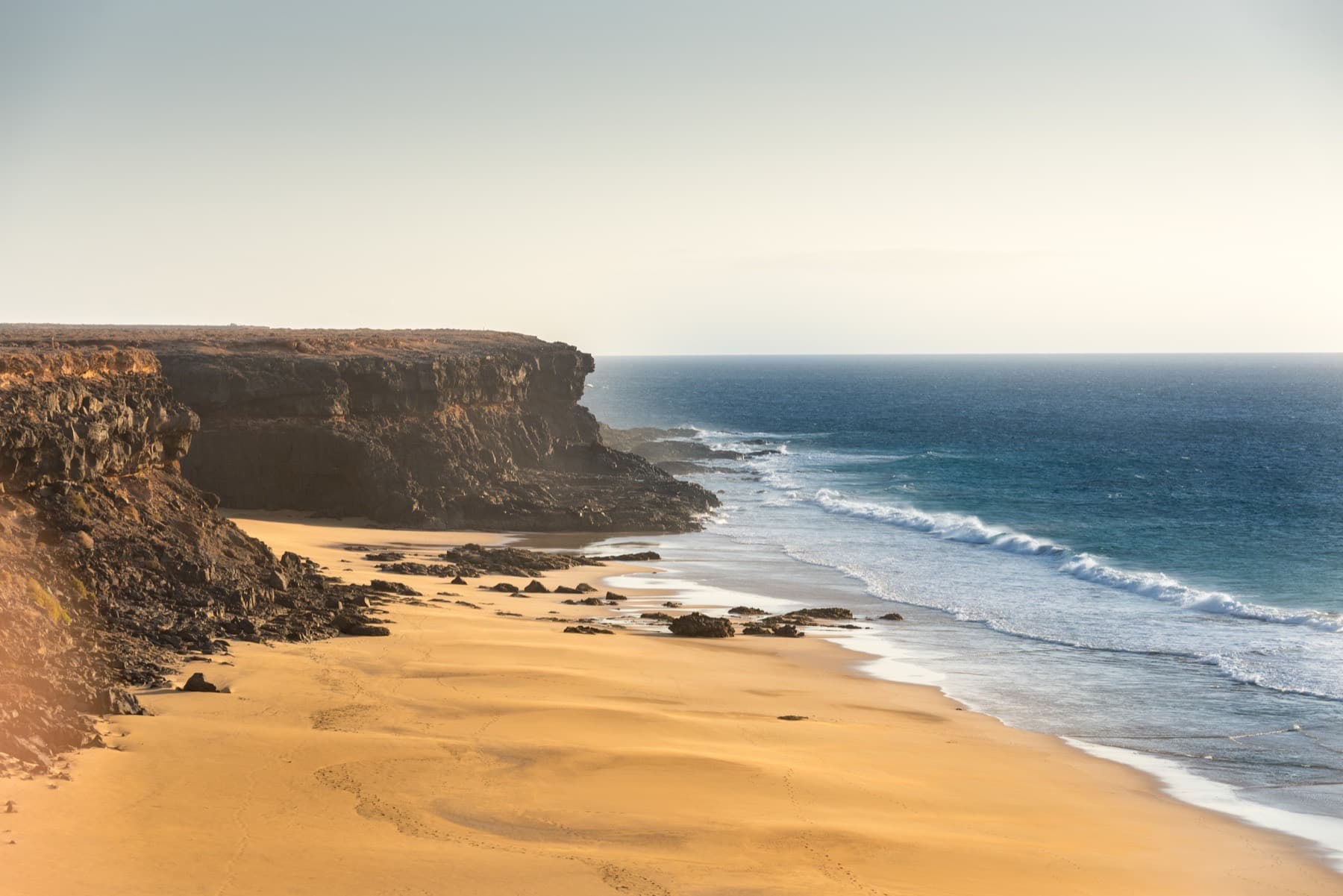 Golden sand beach below dark cliffs, with waves rolling in under a pale sky